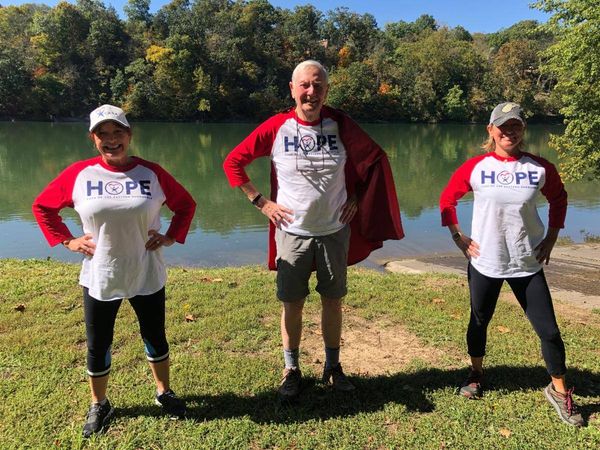 Three people wearing matching red and white H2O branded shirts pose together by a river on a clear day.