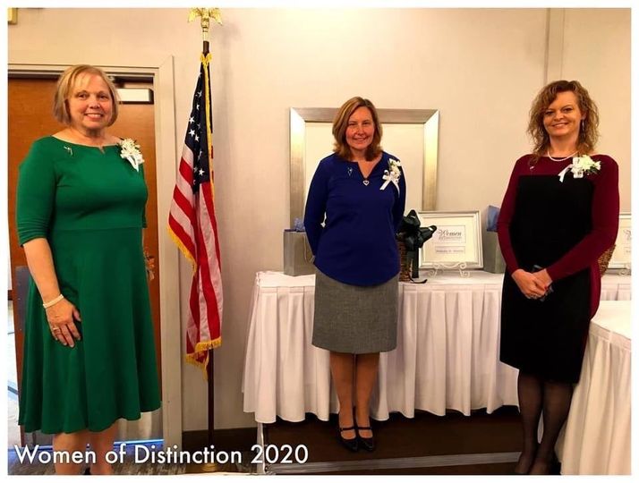 Three women stand formally at an indoor awards ceremony labeled 'Women of Distinction 2020' with an American flag in the background.