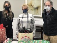 Three people wearing face masks stand behind a table with craft materials and decorative items in an indoor workshop or classroom setting.