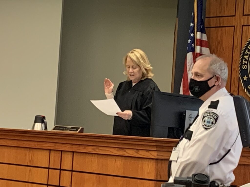A judge and police officer stand at the bench in a courtroom during what appears to be a legal proceeding or hearing.