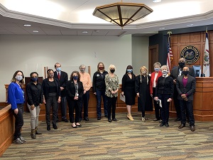 A formal group of approximately 20 people posed together in what appears to be a courtroom or official government building with wood paneling and a decorative ceiling fixture.
