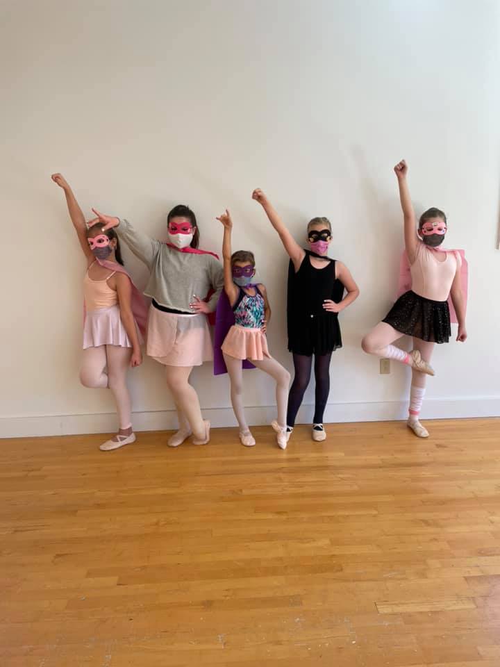 A group of young children in a dance class pose with raised arms in a studio with wooden flooring and white walls.
