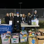 A group of volunteers stand behind a table displaying collected food donations at a 'Change a Child's Story' charity event held at an outdoor sports field.