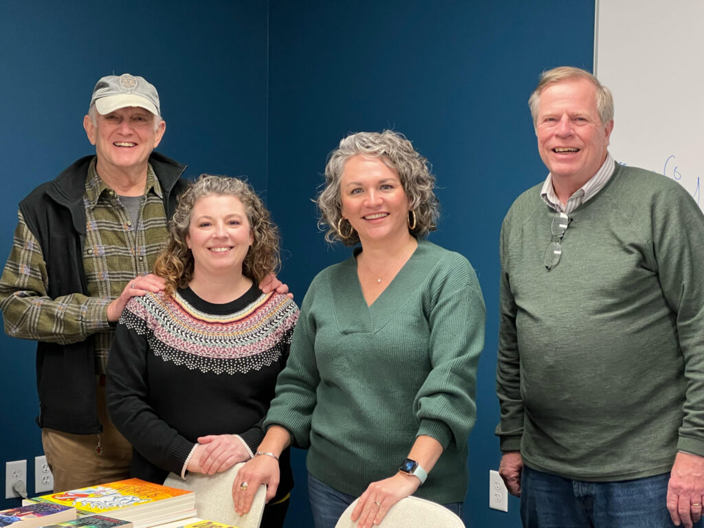 Four adults pose together for a portrait in an indoor workshop or community space with a teal blue wall background.