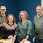 Four adults pose together for a portrait in an indoor workshop or community space with a teal blue wall background.