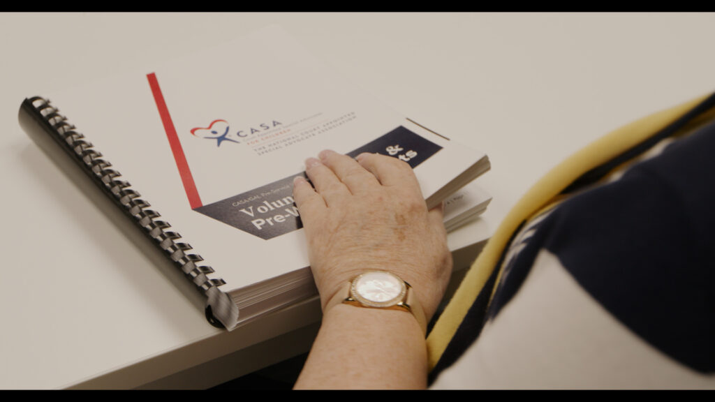 A close-up overhead view of a person's hands writing or working on documents at a desk with a notebook, pen, and wristwatch visible.