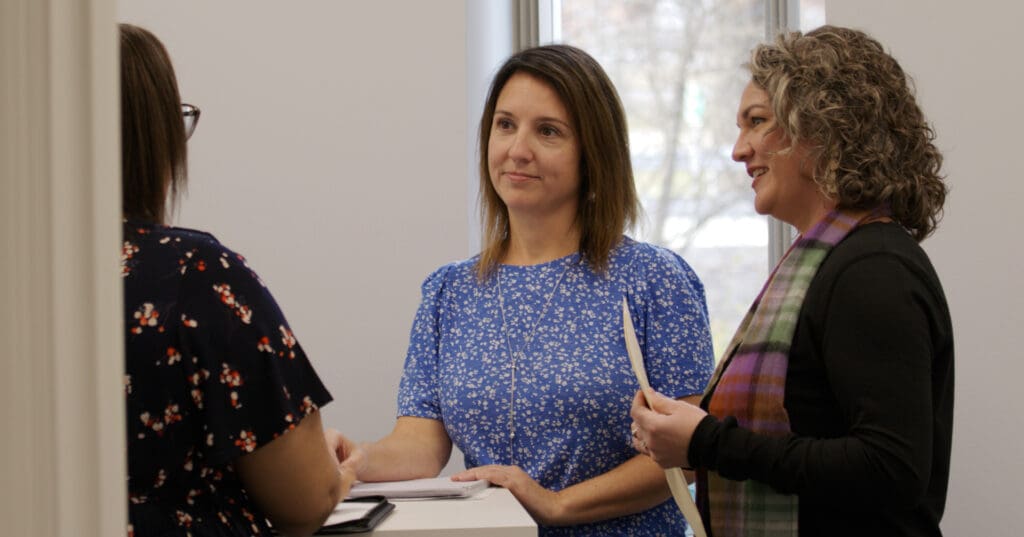 Three women engage in a calm discussion or training session indoors, with one woman in a blue dress as the focal point.