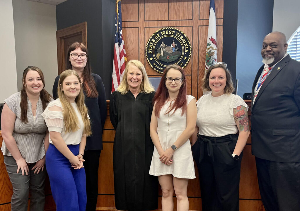 Six women and one man pose formally together in an official government or civic building, standing in front of an American flag and official seal.