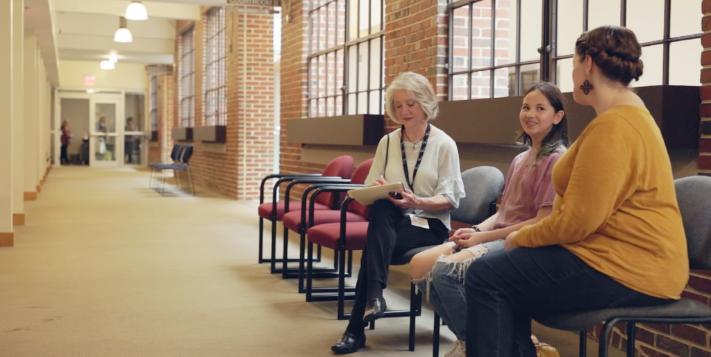 Three people engaged in a calm discussion while seated in folding chairs within a long institutional hallway with brick walls and overhead lighting.