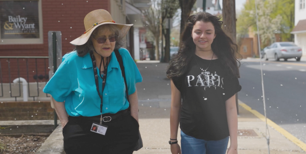 Two people walk down a sunny sidewalk past storefronts, with one wearing a bright turquoise shirt and hat and the other in a dark shirt.