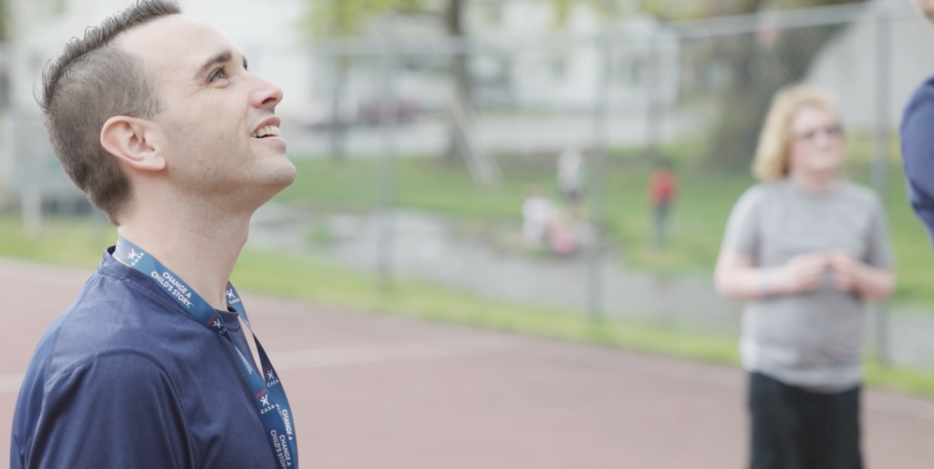 A young man in a navy polo shirt stands in profile at a tennis court facility, looking upward with a calm, contemplative expression.