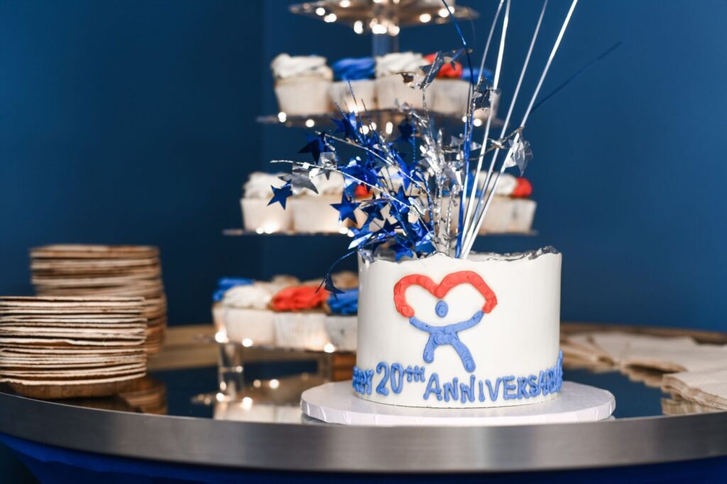 A white anniversary cake decorated with a red heart and blue flowers sits on a dark table against a deep blue background, celebrating a 20th anniversary.