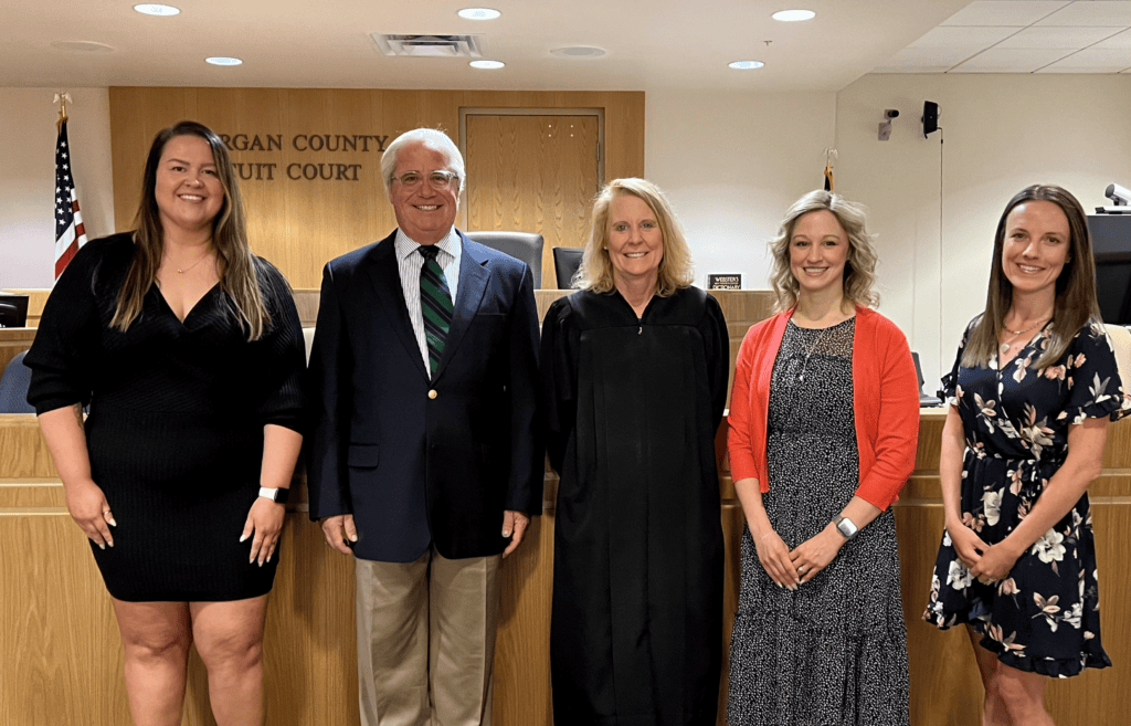 Five professionals stand in a formal group portrait in front of a judge's bench in what appears to be a courthouse or judicial venue.