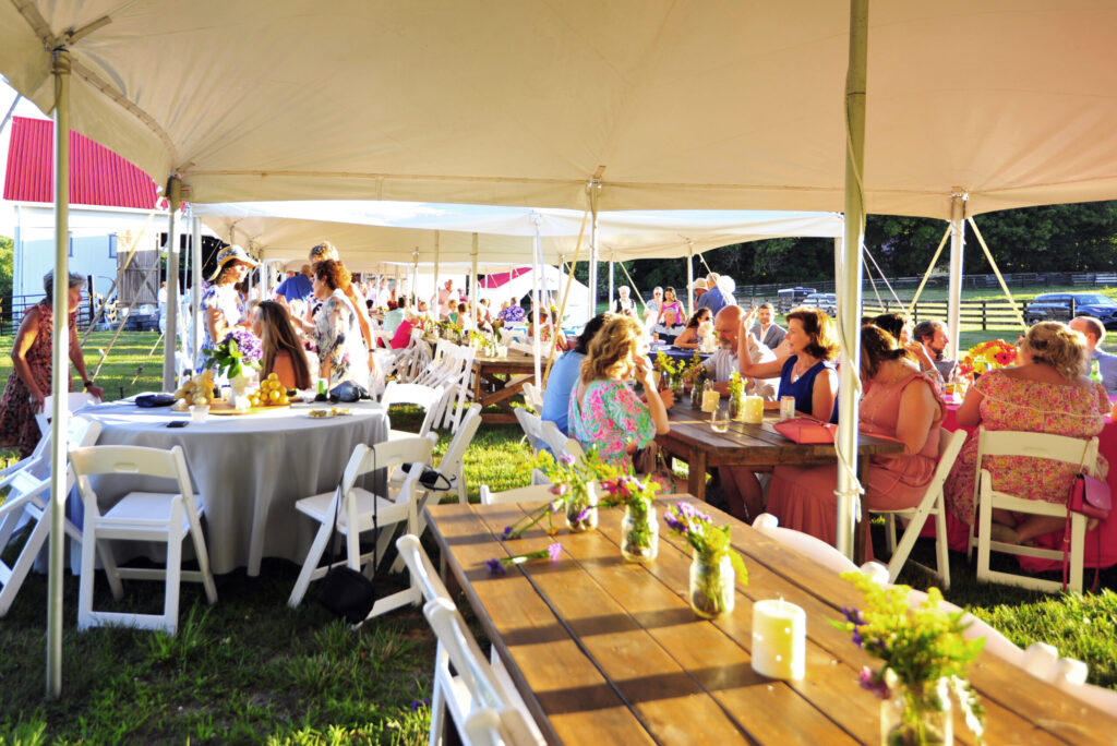 A large outdoor event tent hosts a festive gathering with guests dining at decorated tables under warm golden hour lighting.
