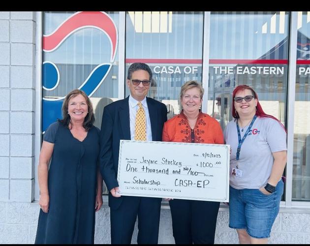 Four professional people stand together holding an oversized ceremonial check for a charitable donation in front of a modern glass building.
