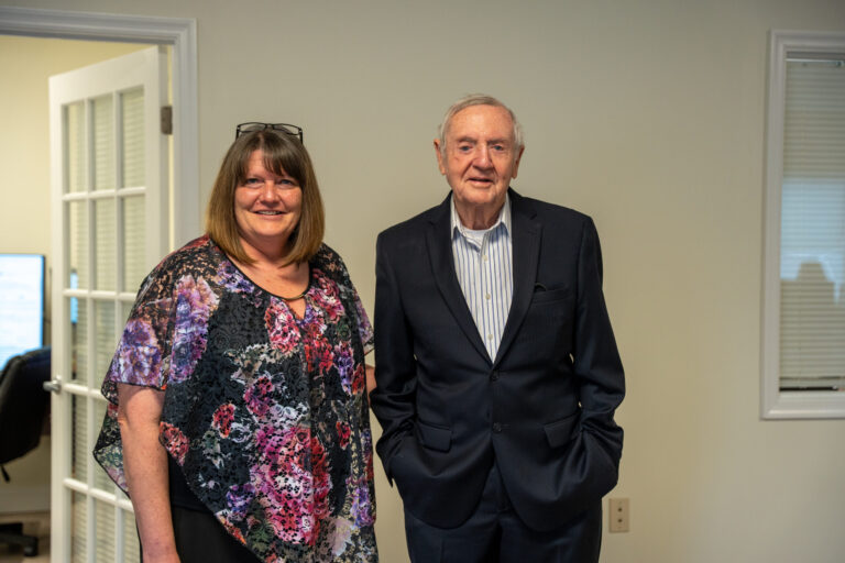 Two professional adults pose together in an indoor office setting with neutral beige walls and white doors.