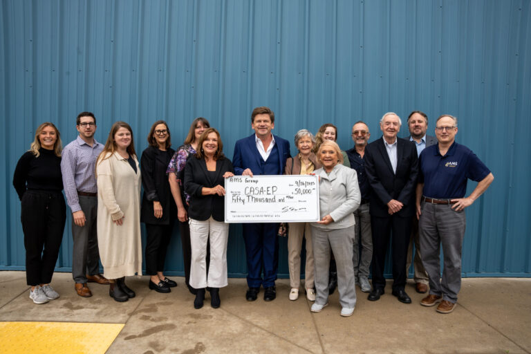 A formal group of approximately 13 people pose together while holding a large ceremonial check in front of an industrial blue corrugated metal wall.