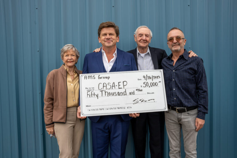 Four people hold an oversized ceremonial check against a blue corrugated metal wall, appearing to celebrate a significant donation or award.