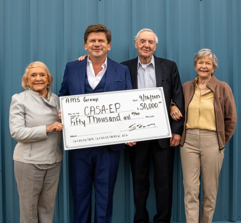 Four people hold an oversized charity check for fifty thousand dollars in front of a blue corrugated metal wall.