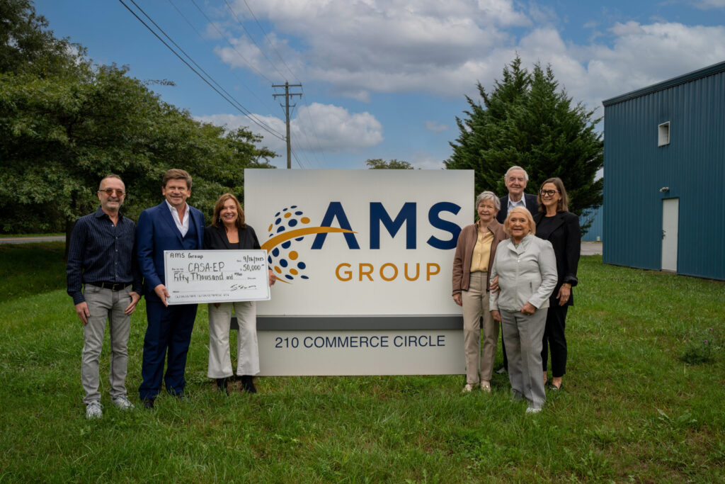 A professional group of eight people pose together outdoors holding a large check in front of an AMS Group sign, with a commercial building and utility infrastructure visible in the background.