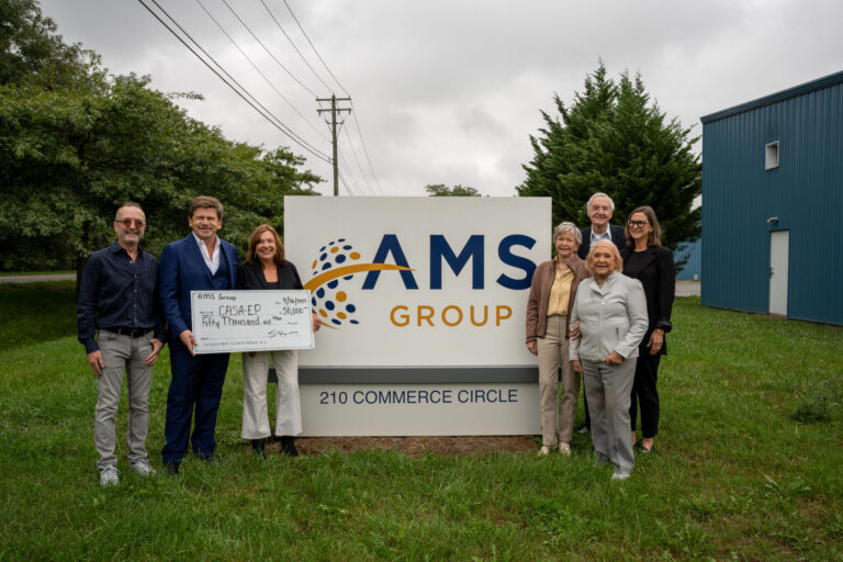 A professional group of eight people pose together with an AMS Group sign in front of a blue commercial building on a grassy field.
