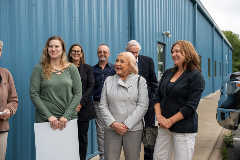 A group of seven people standing in front of a blue corrugated metal building during daytime, appearing to be at a professional or institutional venue.