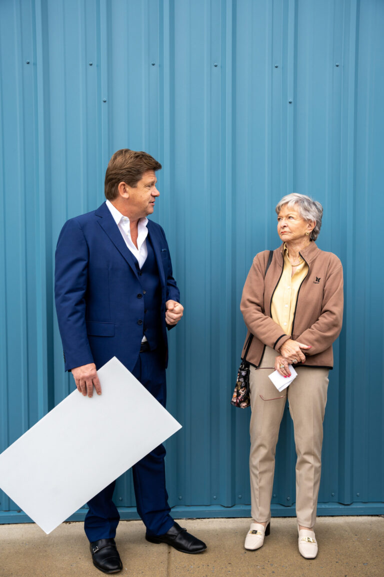 A businessman and businesswoman stand against a bright blue corrugated wall, with the man holding architectural plans during what appears to be a professional consultation.