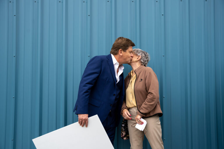 Two business professionals stand against a blue corrugated metal wall while reviewing a blueprint, appearing engaged in a collaborative project discussion.
