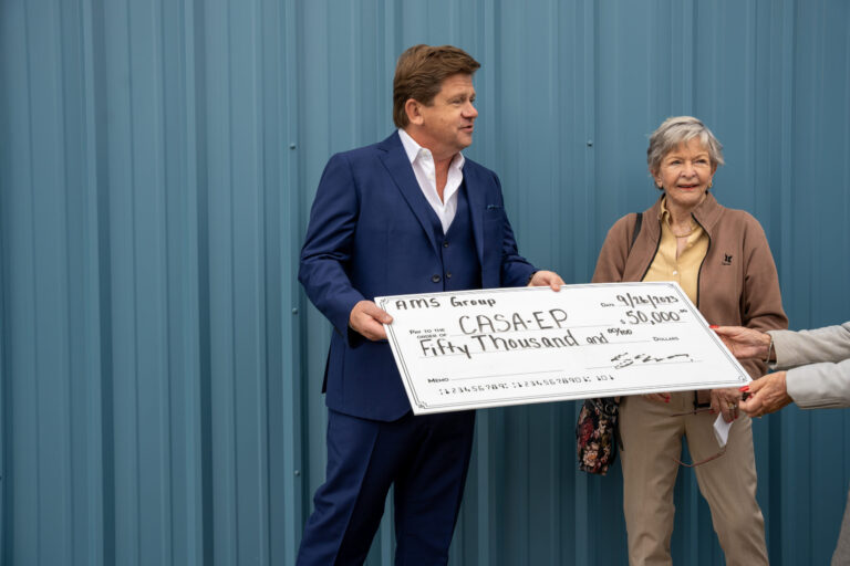 A man in a dark suit and woman present a large ceremonial check against a blue corrugated metal backdrop.