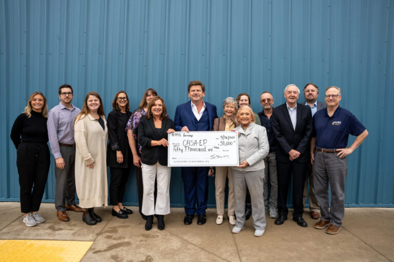 A group of formally dressed individuals stands in front of a corrugated metal wall holding a large ceremonial check, celebrating what appears to be a significant fundraising or donation achievement.
