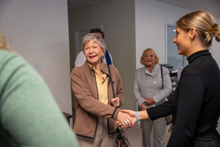 Two professional women shake hands in a modern indoor hallway while other attendees observe in the background during what appears to be a formal meeting or workshop event.