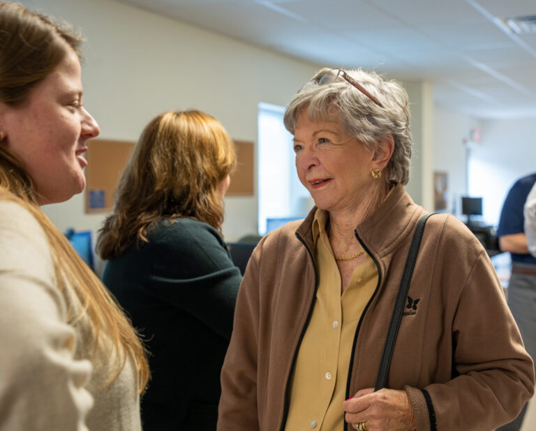 A woman in a brown cardigan engages in conversation at an indoor workshop or community event with other attendees visible in the background.