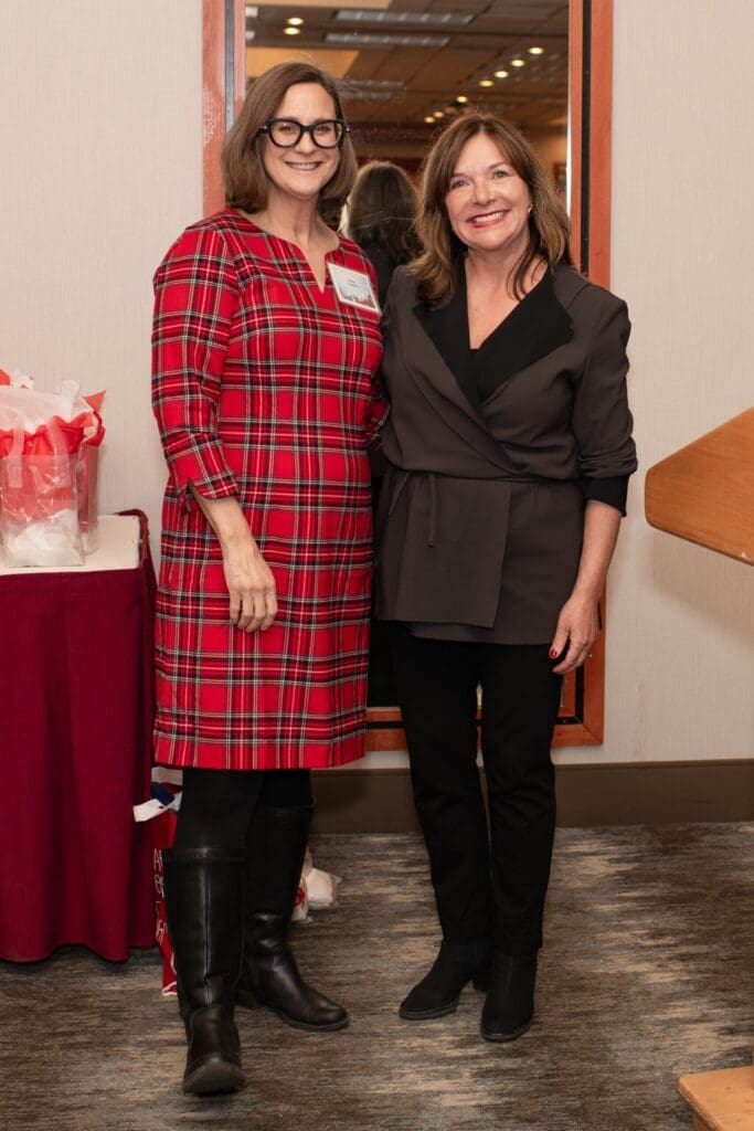 Two women stand together in an indoor venue, one wearing a red plaid dress and the other in a black blazer, posing for a portrait photograph.