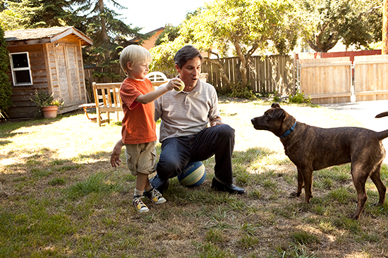 A man crouches down with a young child and a brown dog in a sunny backyard setting surrounded by wooden structures.
