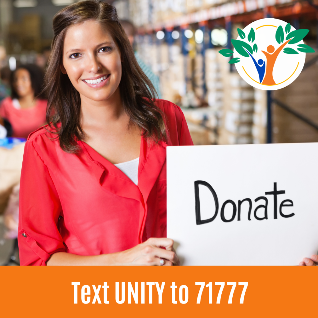 A smiling woman in a red shirt holds a donate sign at what appears to be a charity fundraising event in a shopping venue.