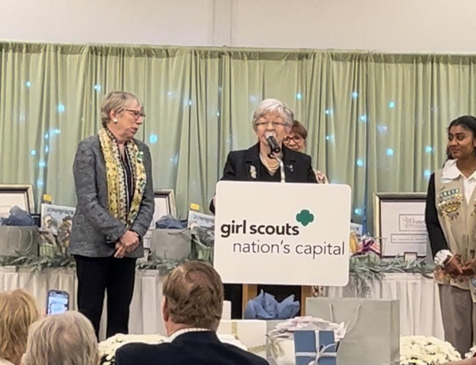 Girl Scouts officials and participants at a formal event or conference with a podium presentation and Girl Scouts banner indicating a nation's capital gathering.