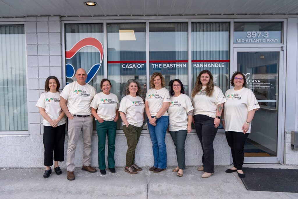 A group of nine people wearing matching light-colored t-shirts stand in a line in front of a storefront with signage.