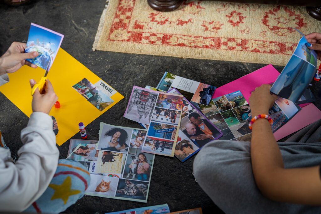 Two people sit on a floor looking at and organizing printed photographs spread around a yellow container during an indoor photo-sharing or organizing session.
