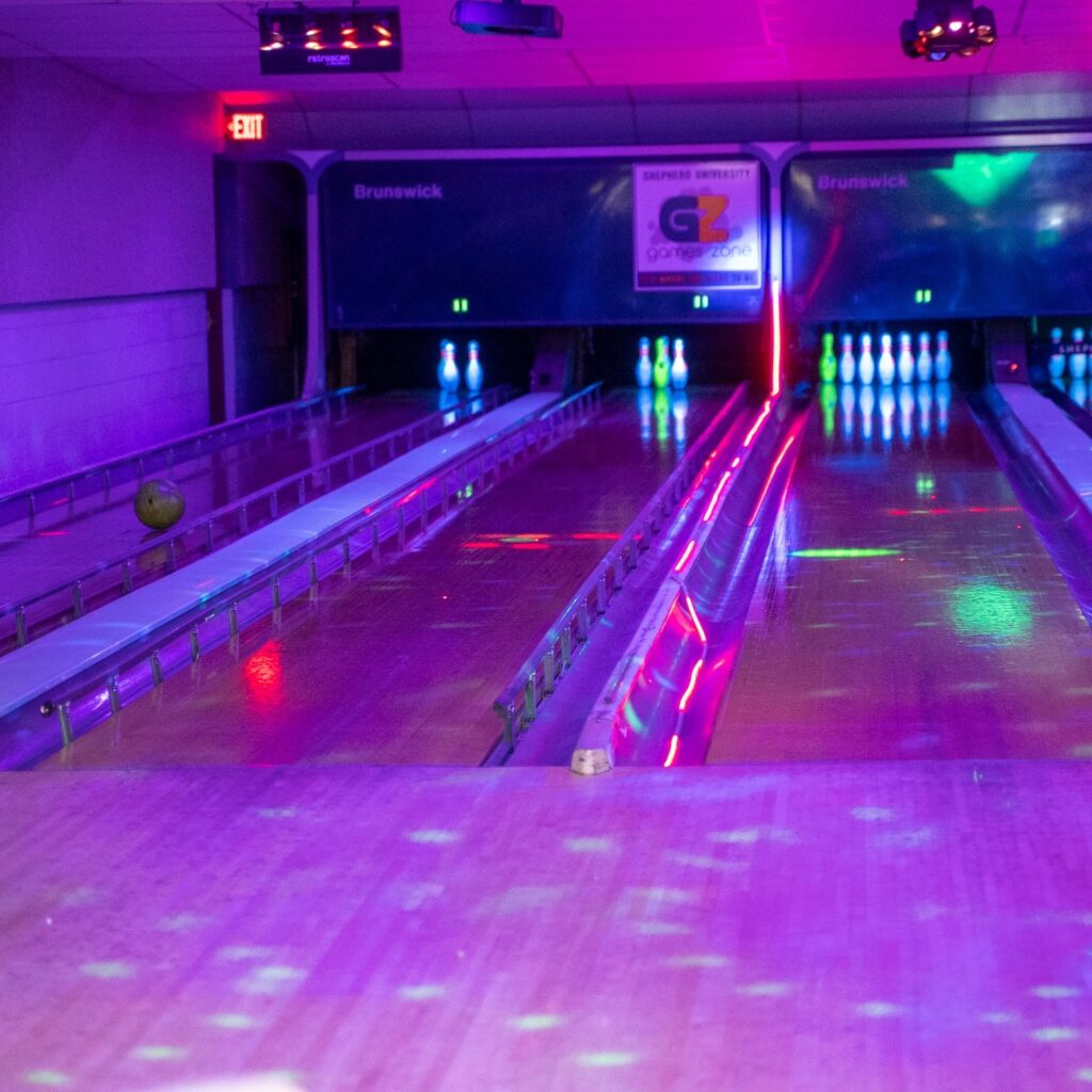 A brightly lit neon bowling alley with glowing purple and pink lighting illuminating empty lanes and white pins.
