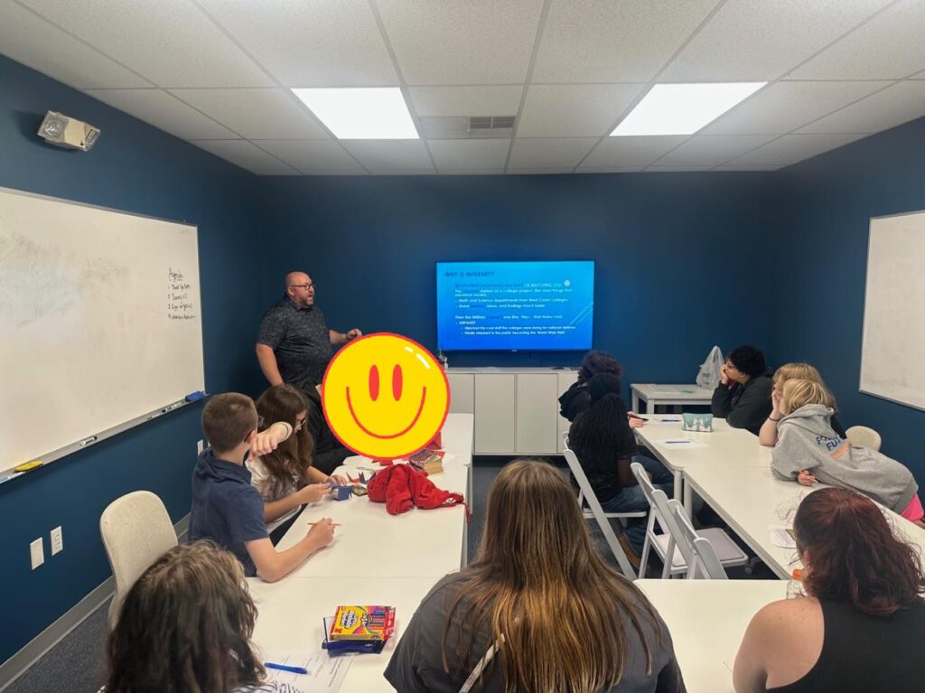 A group of participants attend an indoor workshop or training session in a blue-walled classroom with a presenter, whiteboard, and digital display.