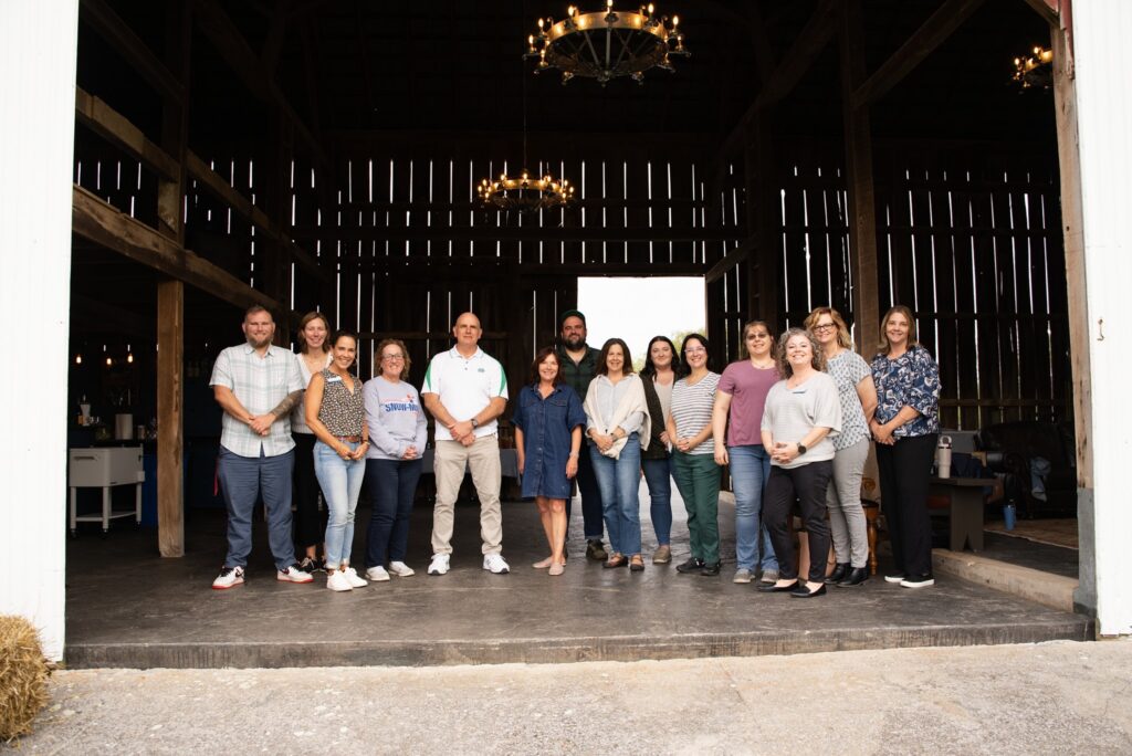 A group of approximately 15-20 people pose together in front of a rustic barn interior with wooden slats and ambient lighting.