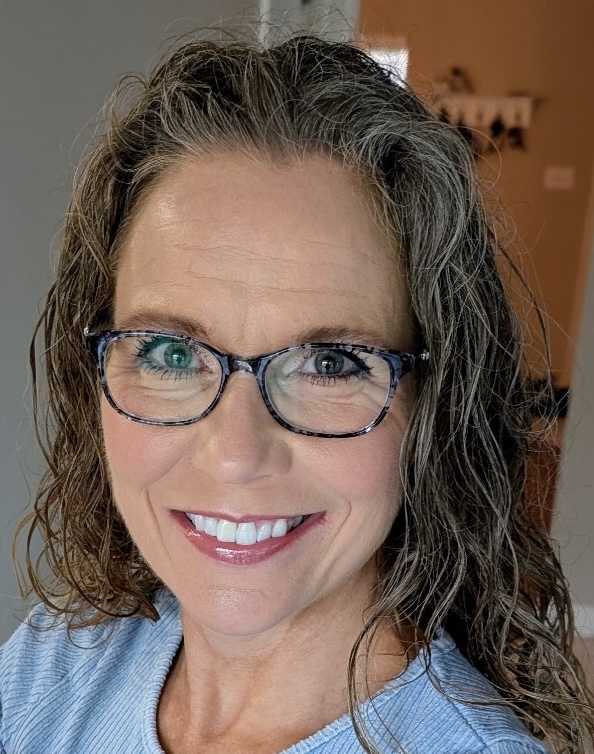 A professional headshot portrait of a smiling woman wearing glasses and a light blue shirt against a neutral indoor background.