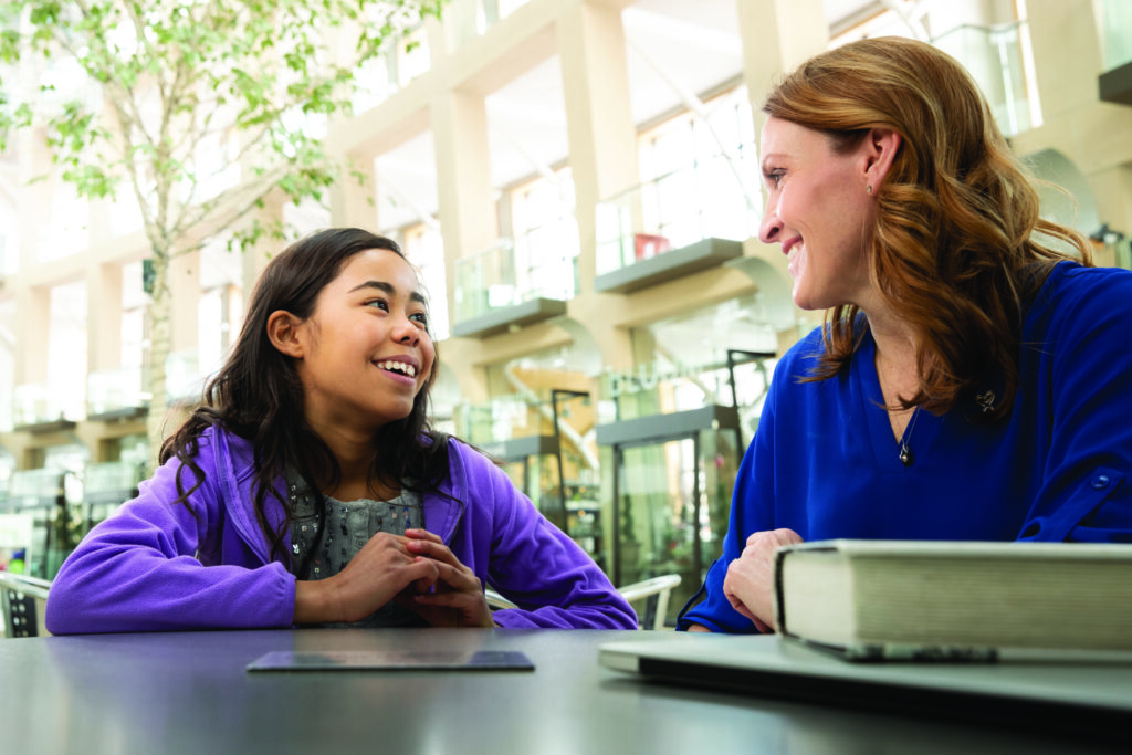 A mentor or teacher in a blue blazer discusses coursework with a student wearing purple in a bright, naturally-lit outdoor classroom setting.