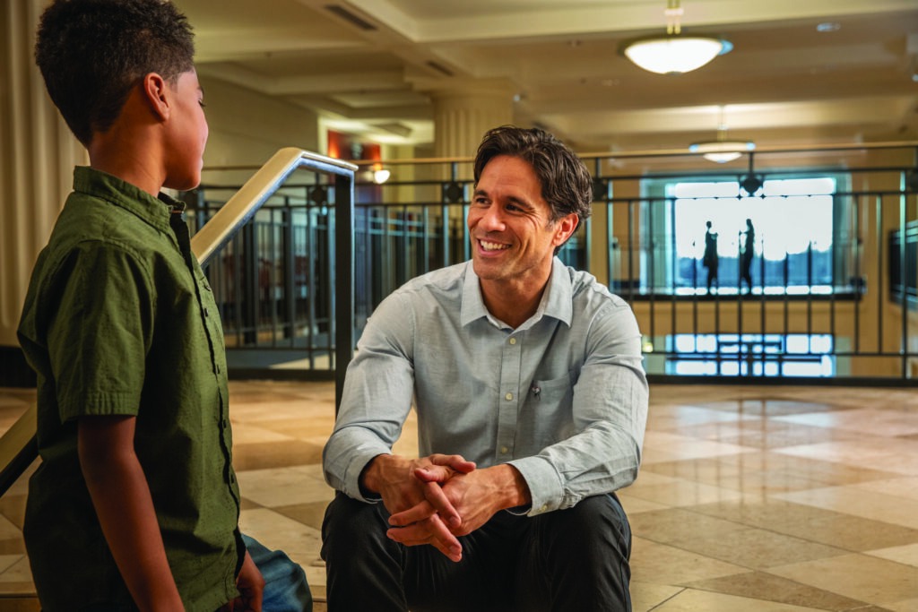 An adult mentor has a one-on-one conversation with a young student in an indoor gymnasium or sports facility, demonstrating engaged teaching or mentorship.