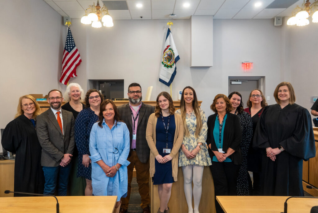 A formal group portrait of approximately 16 people in professional and judicial attire standing in a courthouse or government building, flanked by an American flag and official seal.