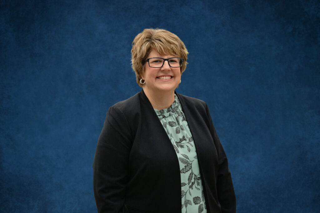 A professional woman wearing glasses and a dark blazer poses against a solid deep blue background in a formal portrait setting.