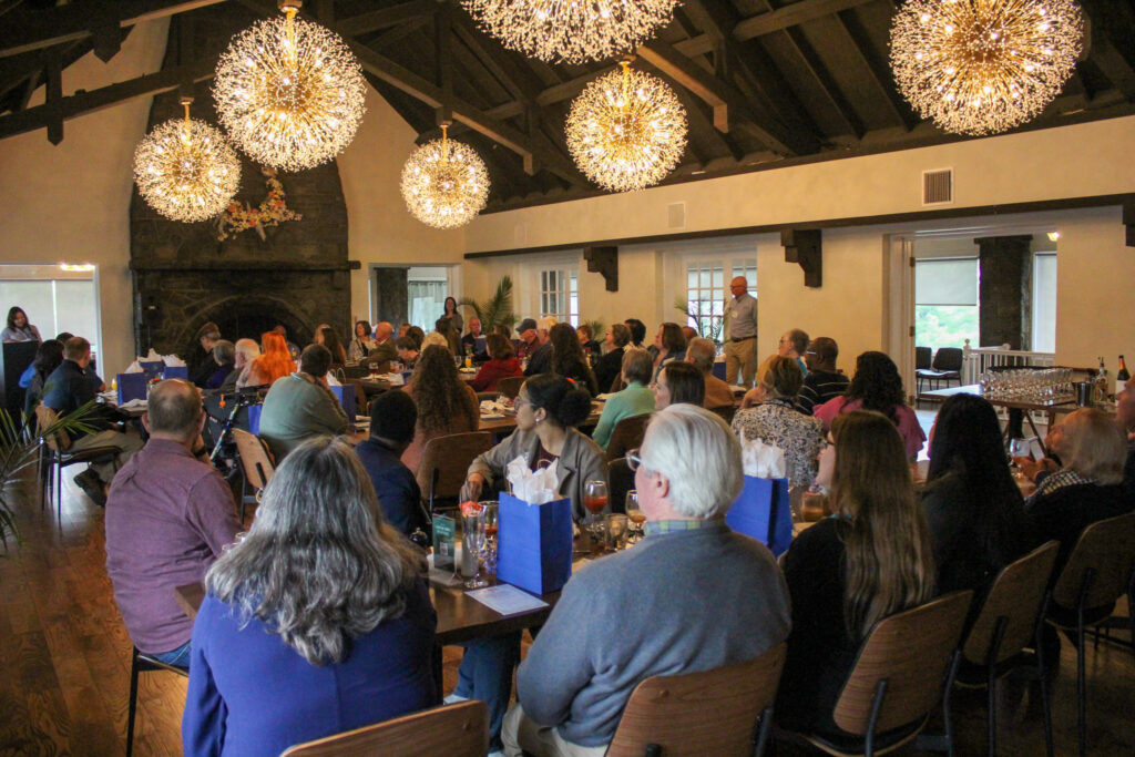 A large indoor crowd gathers in a modern venue with exposed wooden beams and decorative spherical pendant lights casting warm amber light.