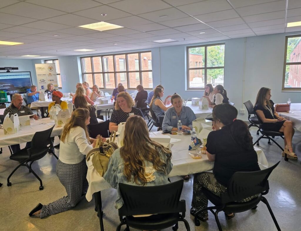 A group of students and educators gather around tables in a bright, light blue classroom during an interactive workshop or collaborative learning session.