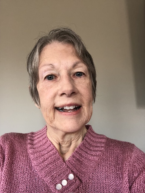 A smiling senior woman wearing a pink knit sweater and pearl necklace poses for a portrait against a neutral beige wall.