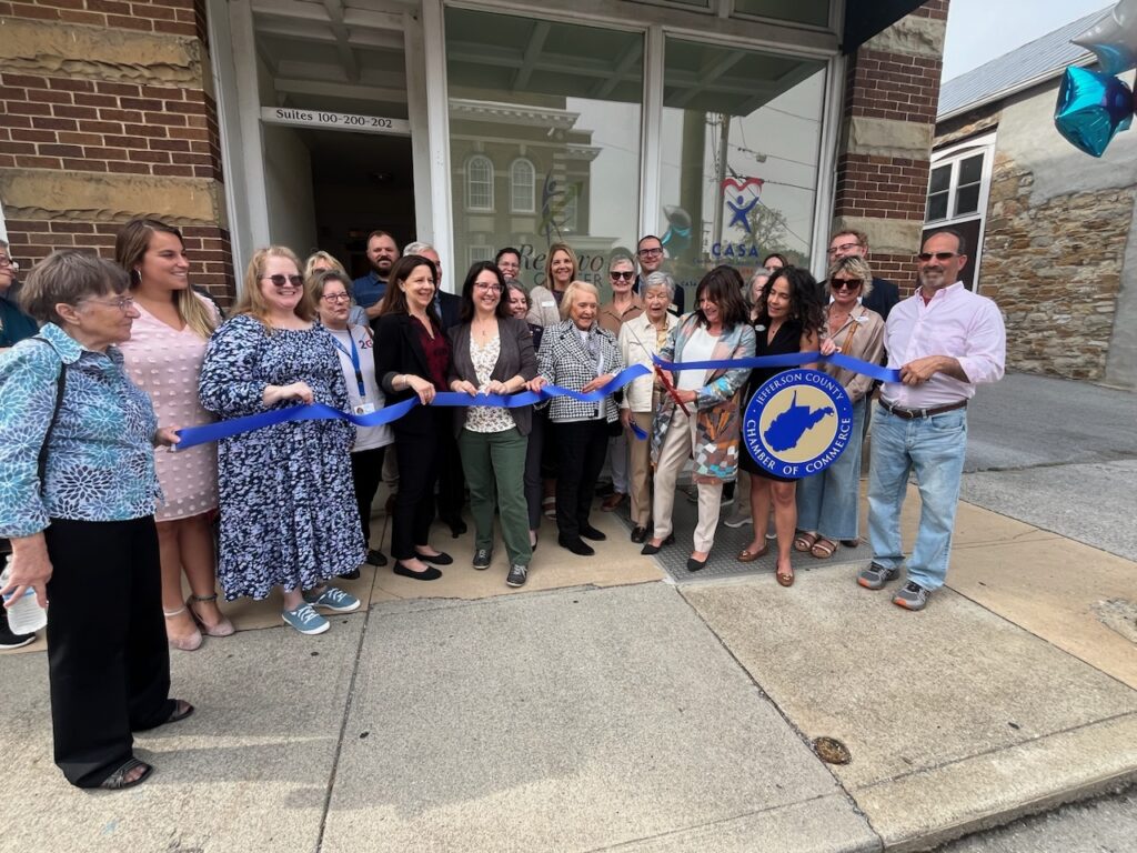 A group of people gather outside a brick building for a ribbon-cutting ceremony, celebrating what appears to be a business opening or grand opening event.